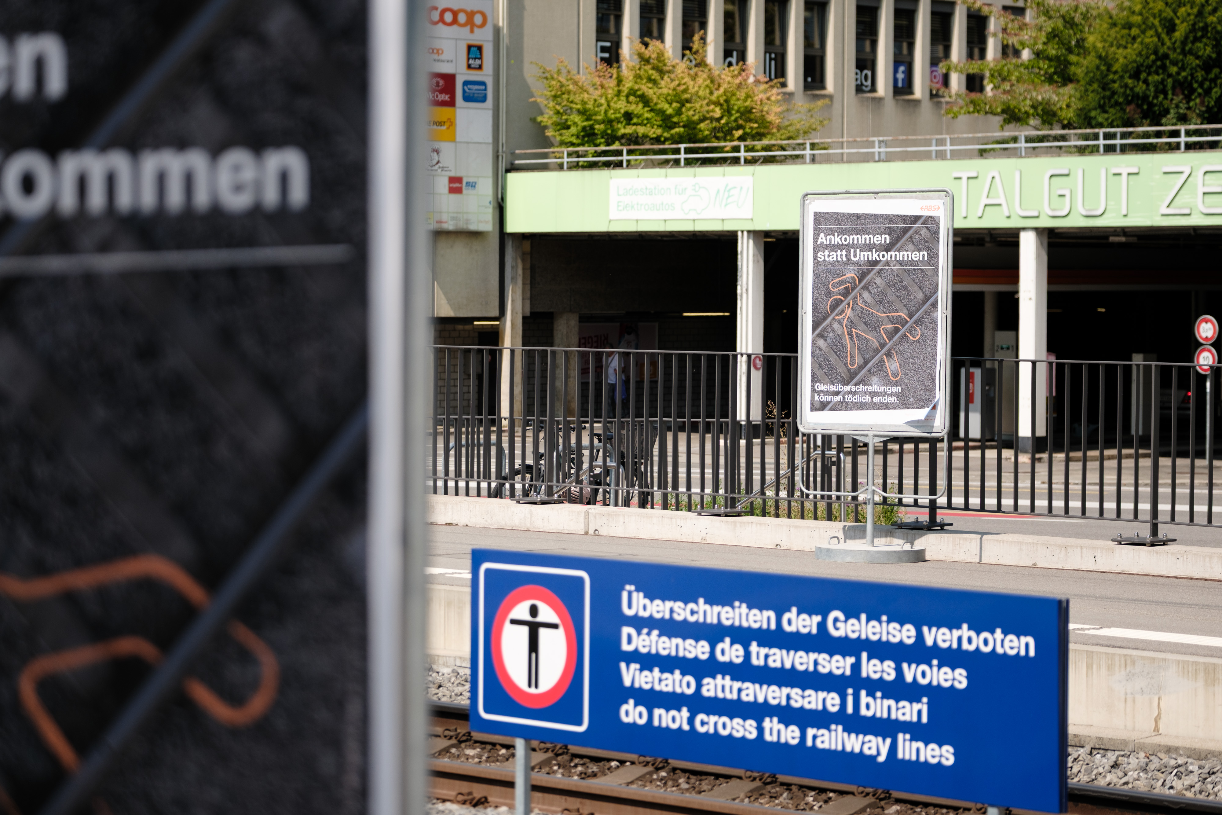 Mit Blick über die Gleise entdeckt man das Kampagnenplakat auf der anderen Seite. Auf der linken Seite ist es ebenfalls angeschnitten. Dazwischen ist das permanente, blaue Schild «Überschreiten der Gleise verboten» zwischen den Gleisen sichtbar.